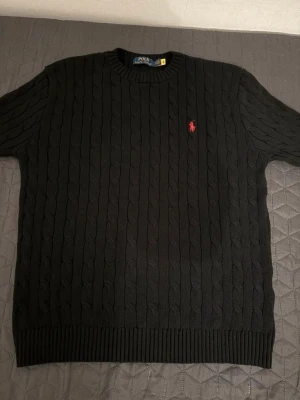 Svart stickad tröja från Polo Ralph Lauren - Svart kabelstickad tröja från Polo Ralph Lauren med rund halsringning och röd broderad logga på bröstet. Tröjan har ribbade muddar och är långärmad, perfekt för kyligare dagar. Klassisk och stilren design som funkar till det mesta.