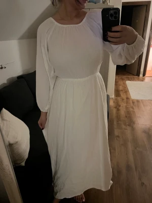 Vit långklänning med snörning i ryggen - Superfin vit långklänning med puffiga långärmar och öppen rygg med snörningsdetaljer. Klänningen har en rundad halsringning och är tillverkad i ett lätt, luftigt material som känns perfekt för varma dagar. Snygg passform med markerad midja och flowy kjol.