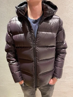 Snoot jacka(ny) - Säljer en svart pufferjacka med stor huva och dragkedja framtill. Jackan har en glansig finish och breda, quiltade paneler som ger en riktigt fet look. Perfekt för kalla dagar när du vill hålla värmen och ändå se stilren ut.