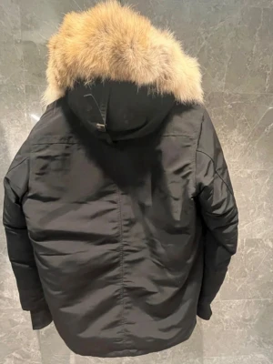 Svart vinterjacka från Canada Goose - Säljer en svart Canada Goose vinterjacka med stor luva och beige pälsdetalj. Jackan har ribbade muddar vid ärmsluten och klassisk patch på ärmen. Perfekt för kalla dagar och riktigt snygg till streetwear-stilen.