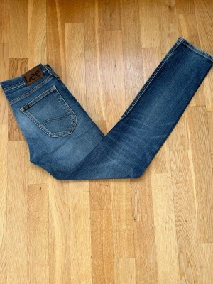 Blå Lee Luke jeans W27 L32 - Säljer ett par blå Lee Luke jeans med klassisk femficksdesign och snygga slitningar. Jeansen har normal passform och raka ben, med Lee-lapp bak i midjan. Tillverkade i slitstarkt denimtyg som ger en tidlös look.