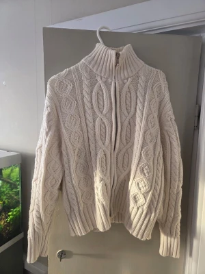 Vit kabelstickad kofta  - Säljer en vit kabelstickad kofta med dragkedja framtill. Tröjan har ett snyggt flätmönster över hela och ribbade muddar vid ärmslut och nederkant. Perfekt för kyliga dagar och riktigt mysig att bära. Tröjan är stl L men den är mindre I storleken så skulle säga att den passar någon som har stl S-M. Pris kan självklart diskuteras😊
