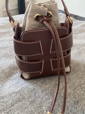 Brun handväska från H&M - Säljer en snygg bucket bag i brunt skinn med vita sömmar och detaljer i guld. Väskan har en beige innerpåse i bomull med snörning upptill och axelrem. Perfekt storlek för det viktigaste och ger en cool, modern look.