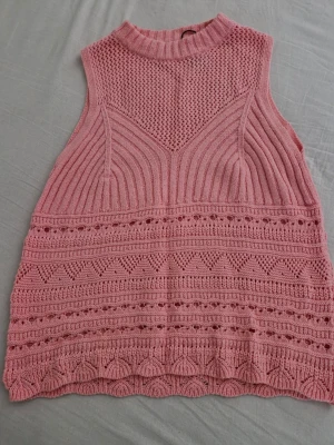 Rosa stickad ärmlös topp från Zara - Säljer en rosa stickad topp från Zara i storlek L. Toppen är ärmlös med hög hals och har ett detaljerat mönster med olika stickade strukturer och hålmönster. Perfekt för att ge din outfit en söt och trendig touch.