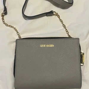 Grå axelväska från Steve Madden - Snygg grå axelväska från Steve Madden med guldfärgade detaljer och kedja. Väskan har en stilren, fyrkantig form och dragkedja upptill. Insidan har flera fack och kortplatser, perfekt för att hålla ordning på dina grejer. Materialet är slätt och känns lyxigt.