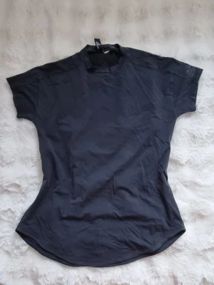 Svart Adidas t-shirt med meshdetalj - Säljer en svart t-shirt från Adidas med klassisk logga på ärmen. T-shirten har korta ärmar, rund hals och meshpanel med små hål på ryggen för extra ventilation. Perfekt för dig som gillar sportig och clean stil.