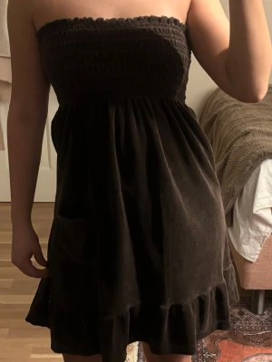 Brun klänning - Säljer en brun axelbandslös kortklänning i mjuk manchester med smock upptill, volangkant nedtill och en praktisk ficka på sidan. Klänningen har en loose passform och är perfekt för dig som gillar bekväma och stilrena plagg.