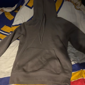 Mörkgrå hoodie med huva och ficka - Säljer en stilren mörkgrå hoodie med stor magficka och huva med snörning. Tröjan har långa ärmar och ribbade muddar vid ärmslut och nederkant. Perfekt för chill dagar eller när du vill ha en enkel look.