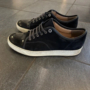Marinblå sneakers från Lanvin med lackad tå - Snygga marinblå sneakers från Lanvin med platt sula och rund tå i blankt lackmaterial. Ovandelen är i mocka och skorna har även marinblåa snören. Insidan är beige och sulan är vit. Perfekt för dig som gillar stilrena sneakers. 