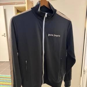 Grå track jacket från Palm Angels XL - Palm Angels track jacket i grått med vita detaljer längs ärmarna och vit dragkedja. Jackan har hög krage, två fickor med dragkedja och logotyp på bröstet. Tillverkad i polyester för en sportig vibe och skön passform. Perfekt för streetwear-stilen.