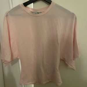 Ljusrosa t-shirt tajt där nere från lager 157, XS - Enkel och stilren ljusrosa t-shirt från lager 157 i storlek XS. T-shirten har rund halsringning och korta ärmar. Tillverkad i mjuk bomull för en skön känsla. Perfekt för en avslappnad look.