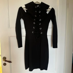 Svart snörad klänning Divided H&M - Svart kort klänning från Divided H&M med långa ärmar och coola cut out-detaljer vid axlarna. Framsidan har snörning med metallöljetter som ger en edgy vibe. Mjuk stretchig polyesterblandning och figurnära passform. Perfekt för dig som gillar statement-plagg.