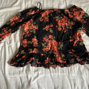 Blommig offshoulder blus från H&M - Säljer en svart blommig offshoulder blus från H&M med röda och gröna rosor. Blusen har långa ärmar och volangdetalj längst ner. Perfekt för dig som gillar romantiska vibes och vill sticka ut med färg och mönster.