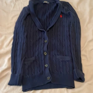 Mörkblå cardigan från Polo Ralph Lauren - Säljer en mörkblå kofta från Polo Ralph Lauren med klassisk kabelstickning, knappar framtill och två fickor. Ikoniska röda loggan broderad på bröstet. Perfekt för lager-på-lager och chill stil. Nypris:2000