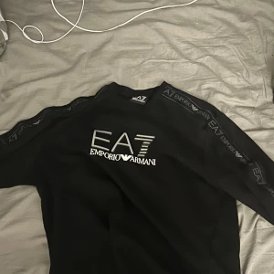 Svart EA7 Emporio Armani sweatshirt - Svart sweatshirt från EA7 Emporio Armani med stor logga framtill och snygga detaljer längs ärmarna. Tröjan har rund hals och lång ärm, perfekt för dig som gillar stilrena och exklusiva märken. Materialet känns mjukt och bekvämt mot huden.
