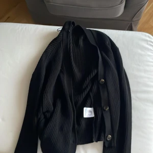 Svart ribbad kofta med knappar - Snygg svart kofta med ribbad struktur och v-ringad hals. Koftan har knappar framtill och är långärmad, vilket gör den enkel att styla. Materialet känns mjukt och stretchigt, perfekt för lager-på-lager. En klassisk och stilren tröja som funkar till många outfits.