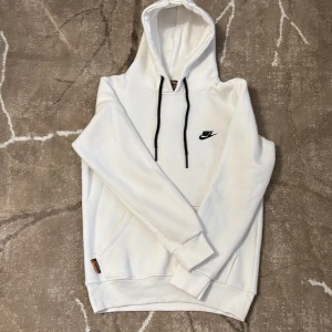 Vit hoodie från Nike med svart logga - Vit hoodie från Nike med broderad svart logga på bröstet och svarta snören i huvan. Tröjan har en klassisk känguruficka framtill och ribbade muddar vid ärmslut och nederkant. Perfekt för chill dagar och streetwear-stil.