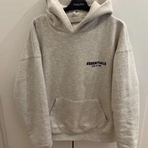  Essentials Fear of God hoodie - Snygg ljusgrå hoodie från Essentials Fear of God med svart logotyptryck både fram och bak. Tröjan har huva, känguruficka och en loose passform som ger en chill vibe. Perfekt för dig som gillar streetwear och vill ha något bekvämt och stilrent. 