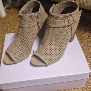 Madden Girl - Beige sandaletter med klack  - Snygga beige sandaletter från Madden Girl med hög klack och öppen tå. Skorna är tillverkade i mocka med perforerat mönster och har dekorativt bälte med nitar runt ankeln. Dragkedja på sidan för enkel påtagning. Perfekt för dig som gillar trendiga detaljer. Sandaletter är i mycket bra skick. Användes bara ett par gånger och har inga defekter.