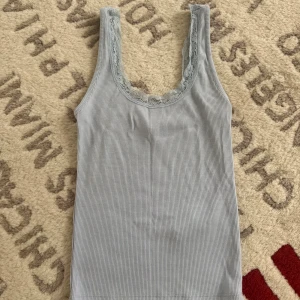 Ljusblå ribbad linnetopp från Gina Tricot - Supersöt ljusblå linnetopp från Gina Tricot i ribbad bomull med spetskant runt halsringningen. Toppen är ärmlös och har en smickrande, lite djupare urringning. Perfekt att styla med jeans eller kjol för en chill look.