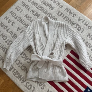 Beige stickad kofta med knyt från h&m - Supermysig beige stickad kofta från h&m med grov ribbad struktur och breda muddar. Koftan har ett snyggt knytband i midjan och långärmade ärmar. Perfekt för lager-på-lager och chill dagar. Passar dig som gillar en relaxed och cozy vibe.