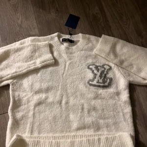 Vit stickad Louis Vuitton tröja S - Exklusiv vit stickad tröja från Louis Vuitton i storlek S men mer som M Tröjan har ett stort grått LV-logo broderat på bröstet, ribbade muddar och rund hals. Mjuk och fluffig känsla, perfekt för dig som gillar lyxiga och stilrena plagg.
