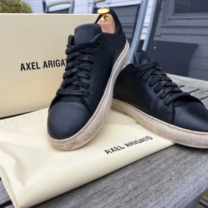 Svarta sneakers från Axel Arigato - Stilrena svarta sneakers från Axel Arigato i skinn med vit platt sula och diskret logga på sidan och hälen. Klassisk rund tå, svarta skosnören och minimalistisk design som passar till allt. Kommer med originalkartong och dustbag.
