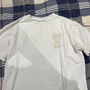 Vit t-shirt med broderat hjärta  - Vit t-shirt i mjuk bomull med korta ärmar och rund hals. På bröstet finns ett broderat hjärta med rosett i samma färg som tröjan, vilket ger en clean och stilren look. Perfekt för dig som gillar enkla detaljer och neutrala färger.