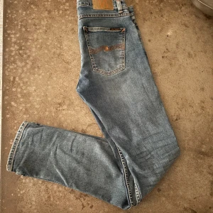 Blå jeans från Nudie Jeans Co, 29/32 - Snygga ljusblå jeans från Nudie Jeans Co med klassisk femficksdesign och orangea sömmar. Jeansen har en rak passform och är tillverkade i mjukt denim med diskret tvättad look. Bakfickan har Nudies ikoniska broderi i orange.