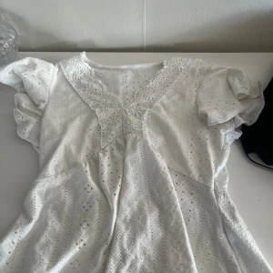 Vit kort klänning med spetsdetaljer - Superfin vit kort klänning med broderade hålmönster och v-ringning med spetsdetaljer. Puffiga korta ärmar och lätt utställd kjol ger en romantisk vibe. Perfekt för varma dagar och somriga tillfällen.