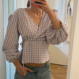 Ljuslila rutig omlottblus med puffärm - Säljer en superfin ljuslila och vit rutig omlottblus med puffiga trekvartsärmar och knyt i midjan. Blusen har v-ringad hals och croppad passform som ger en fräsch och trendig look. Perfekt för vår och sommar, och är gjord i mjuk bomull.