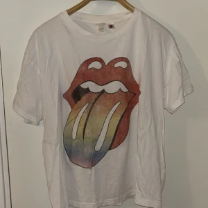 Rolling Stones t-shirt från H&M XL - Vit t-shirt från H&M L.O.G.G. i storlek XL med ikoniskt Rolling Stones-tryck framtill och bandets logga i svart på ryggen. Klassisk rund hals och korta ärmar. Materialet är mjuk bomull, perfekt för dig som gillar bandmerch och vill sticka ut med en färgglad graphic tee.