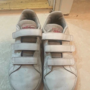 Vita Adidas sneakers med kardborre - Säljer ett par klassiska vita Adidas sneakers för barn med tre kardborreband och perforerade detaljer på sidorna. Skorna har röd hälflik med Adidas-logga och är tillverkade i syntetmaterial. Perfekta för en sportig och clean look.