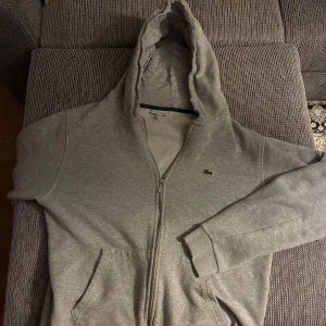 Lacoste Zip hoodie - Hej, säljer nu min Lacoste ZipUp i färgen grå som är i nyskick. Använd ett fåtal gånger utan fläckar och slitage. Jag har storlek S och den passade mig men borde även passa XS bra. Kolla storleken på bilden.