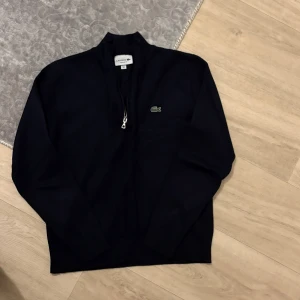 Marinblå stickad tröja från Lacoste - Snygg marinblå stickad tröja från Lacoste med halv dragkedja och klassisk krokodillogga på bröstet. Tröjan har långärmad design och ribbad krage som ger en clean och stilren look. Perfekt för dig som gillar sportig och tidlös stil.