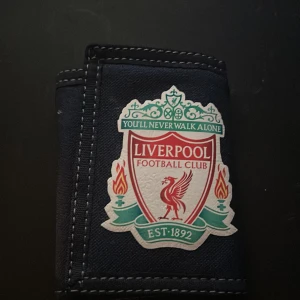 Liverpool FC svart plånbok - Svart plånbok med Liverpool Football Club-logga på framsidan. Plånboken har vita sömmar, flera kortfack, ett fönster för ID och en metallring för nycklar. Materialet är slitstark polyester och designen är klassisk och sportig.