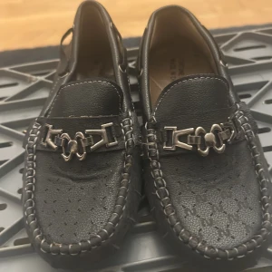 Svarta loafers med metallspänne - Snygga svarta loafers i skinn med dekorativt metallspänne framtill. Skorna har rund tå, perforerat mönster på ovandelen och grovsydda kanter. Yttersulan har gummidetaljer för extra grepp. Perfekta för en stilren och klassisk look.