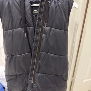 Svart quiltad dunväst från ONLY - Svart dunväst från ONLY med huva och snörning. Västen har quiltad design, dragkedja framtill och är tillverkad i syntetmaterial. Perfekt för lager-på-lager och har en clean, modern look med snygga detaljer på snörningen.