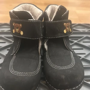 Svarta barnkängor med nallebrodyr - Svarta kängor för barn med vit kontrastsöm och kardborreband dekorerade med broderade nallar. Skorna har en robust sula och är fodrade med mjukt vitt material för extra värme. Perfekta för kyligare dagar och lek utomhus.