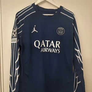 PSG långärmad matchtröja Dembélé #10 - Snygg blå PSG långärmad fotbollströja med vita detaljer, Dri-Fit-material och O. Dembélé #10 på ryggen. Jordan-logga, Qatar Airways-tryck och Champions League-märke på ärmen. Perfekt för dig som älskar fotboll och vill sticka ut på läktaren.