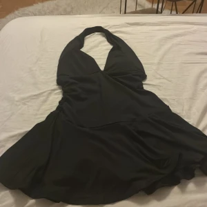 Svart halterneck klänning FB Sister XS - Supersnygg svart kortklänning från FB Sister i storlek XS. Klänningen har halterneck och en djup v-ringning framtill som ger en trendig look. Materialet är mjukt och har ett lätt glansigt fall. Perfekt för dig som gillar stilrena och enkla plagg med lite edge.