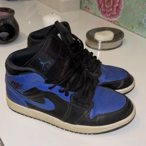 Nike Air Jordan 1 Mid Svart/Blå - Säljer ett par Nike Air Jordan 1 Mid i svart och blått skinn med klassisk vinge-logga på sidan och blå swoosh. Skorna har svart snörning, vit mellansula och svart yttersula. Perfekta för dig som gillar streetwear och vill sticka ut med en ikonisk sneaker.