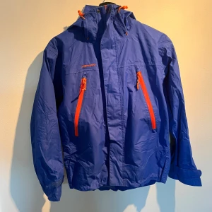 Blå skidjacka från Stormberg - Blå skidjacka från Stormberg med orange dragkedjor och detaljer. Jackan har huva, två snedställda bröstfickor med dragkedja och justerbara ärmslut. Perfekt för blåsiga dagar och outdoor-aktiviteter. Snygg och funktionell design.