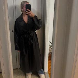 Svart oversized skinn trenchcoat - Säljer en svart trenchcoat i skinnimitation med oversized passform och breda slag. Jackan har bälte i midjan, långa ärmar och djupa fickor. 
