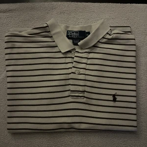 Randig pikétröja från Polo Ralph Lauren - Snygg vit pikétröja med svarta horisontella ränder från Polo Ralph Lauren. Klassisk krage, korta ärmar och knäppning med två knappar framtill. Ikonisk broderad logga på bröstet. Tillverkad i 100% bomull för skön och luftig känsla.