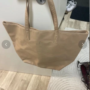 Beige shoppingväska med dragkedja - Stor beige shoppingväska med två långa handtag och praktisk dragkedja på utsidan. Väskan är tillverkad i syntetmaterial och har ett rymligt innerfack med extra ficka med dragkedja. Perfekt för att bära med sig allt man behöver under dagen.