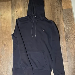 Svart hoodie från GANT med huva - Marinblå hoodie från GANT i storlek S med klassisk logga broderad på bröstet. Tröjan har huva med snörning, magficka och ribbade muddar. Materialet är mjuk bomull och polyester, perfekt för chill dagar. Snygg och enkel design som funkar till jeans eller joggers.