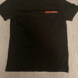 Svart t shirt med rött tryck - Svart t-shirt med Prada tryck. Kan inte garantera äkthet därav priset & litet hål i fram som syns på första bilden, kan nog lätt sys igen snabbt men vill bli av med den. Skriv vid frågor!