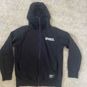 Svart hoodie från Hoodrich med meshdetaljer - Svart hoodie från Hoodrich med vit logga på bröstet och tryck på ryggen. Tröjan har meshdetaljer vid huvan, hel dragkedja framtill och fickor med dragkedja. Långärmad och med ribbade muddar för extra komfort. Perfekt för streetwear-stilen.