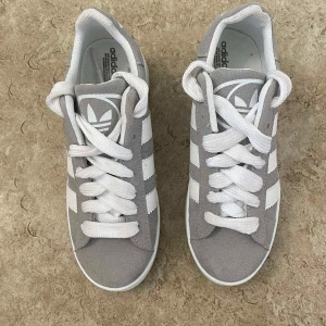 Adidas Campus grå sneakers - Snygga Adidas Campus sneakers i ljusgrå mocka med klassiska vita ränder på sidorna och vit platt sula. Skorna har rund tå, vit snörning och Adidas logga på plösen. Perfekt för dig som gillar stilrena och tidlösa sneakers.
